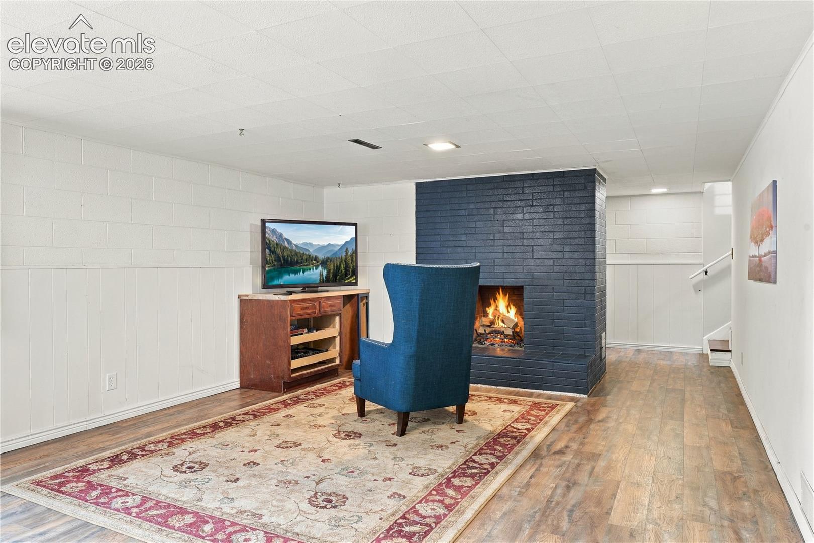 Painted brick fireplace with a wide hearth, wood-finish flooring, white block-patterned walls, and recessed ceiling lighting