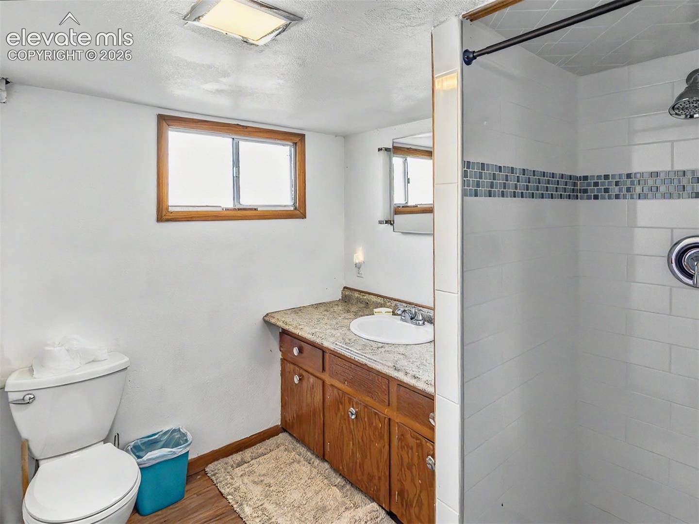 Basement Bathroom featuring a walk-in shower with white subway tile and decorative mosaic band, wood-finish flooring, a vanity with wood cabinetry and a countertop, and a window.