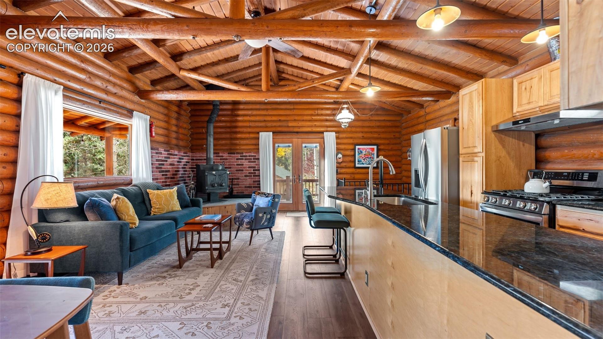 Log-walled interior featuring an exposed timber beam ceiling, wood-burning stove with brick surround, kitchen island with dark stone countertop, and double glass-panel doors