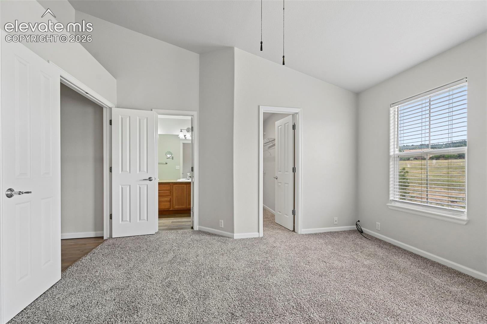 Spacious room featuring vaulted ceilings, light gray carpeting, a large window with horizontal blinds, and a private bath with a wood-finish vanity