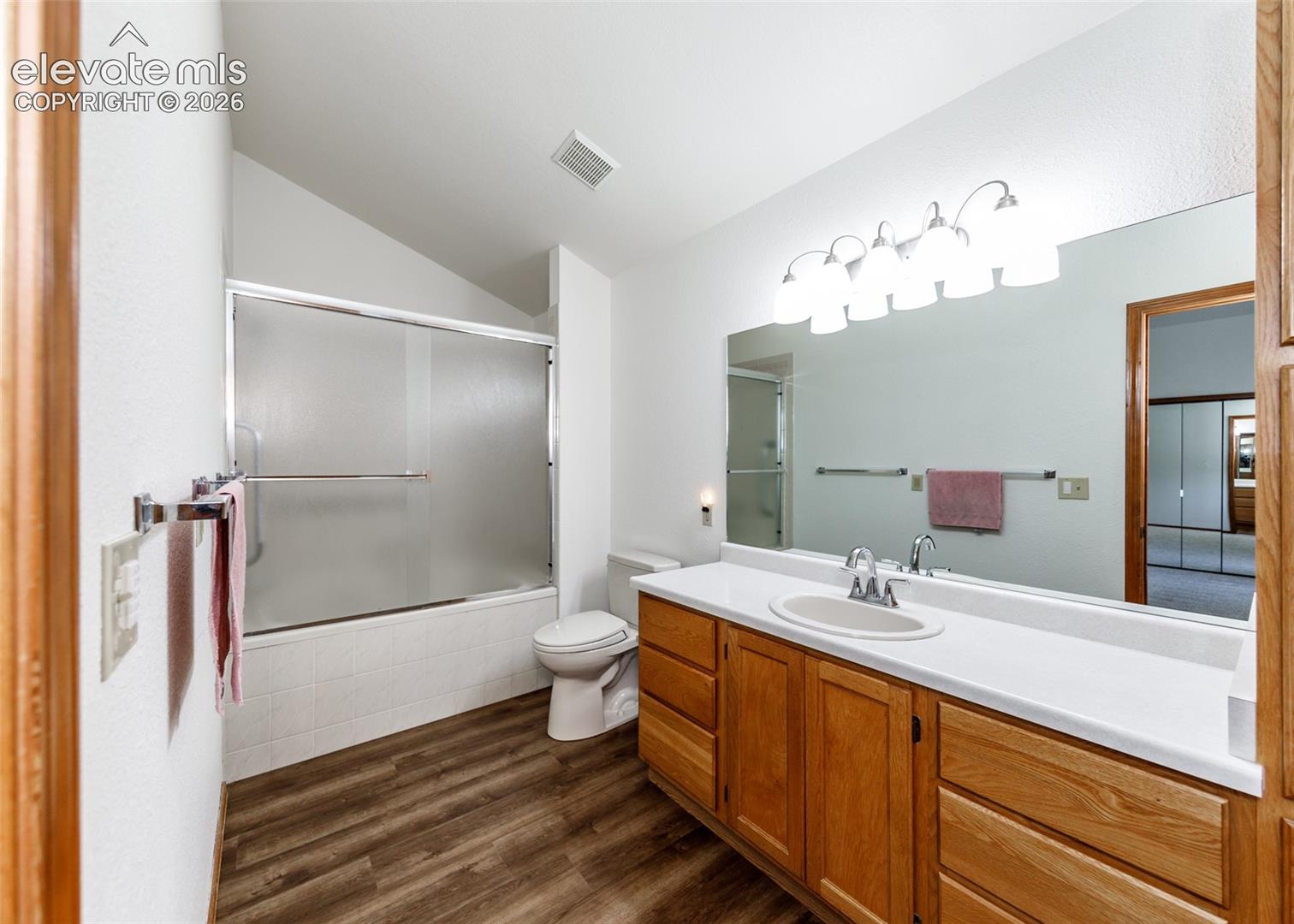 Wood-finish flooring, wood cabinetry, built-in vanity with integrated sink, full-width mirror, and a shower-over-tub with sliding frosted glass doors