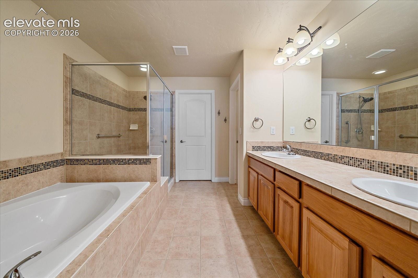 Dual-sink wood vanity with a tile countertop, soaking tub, glass-enclosed shower, and decorative tile backsplash