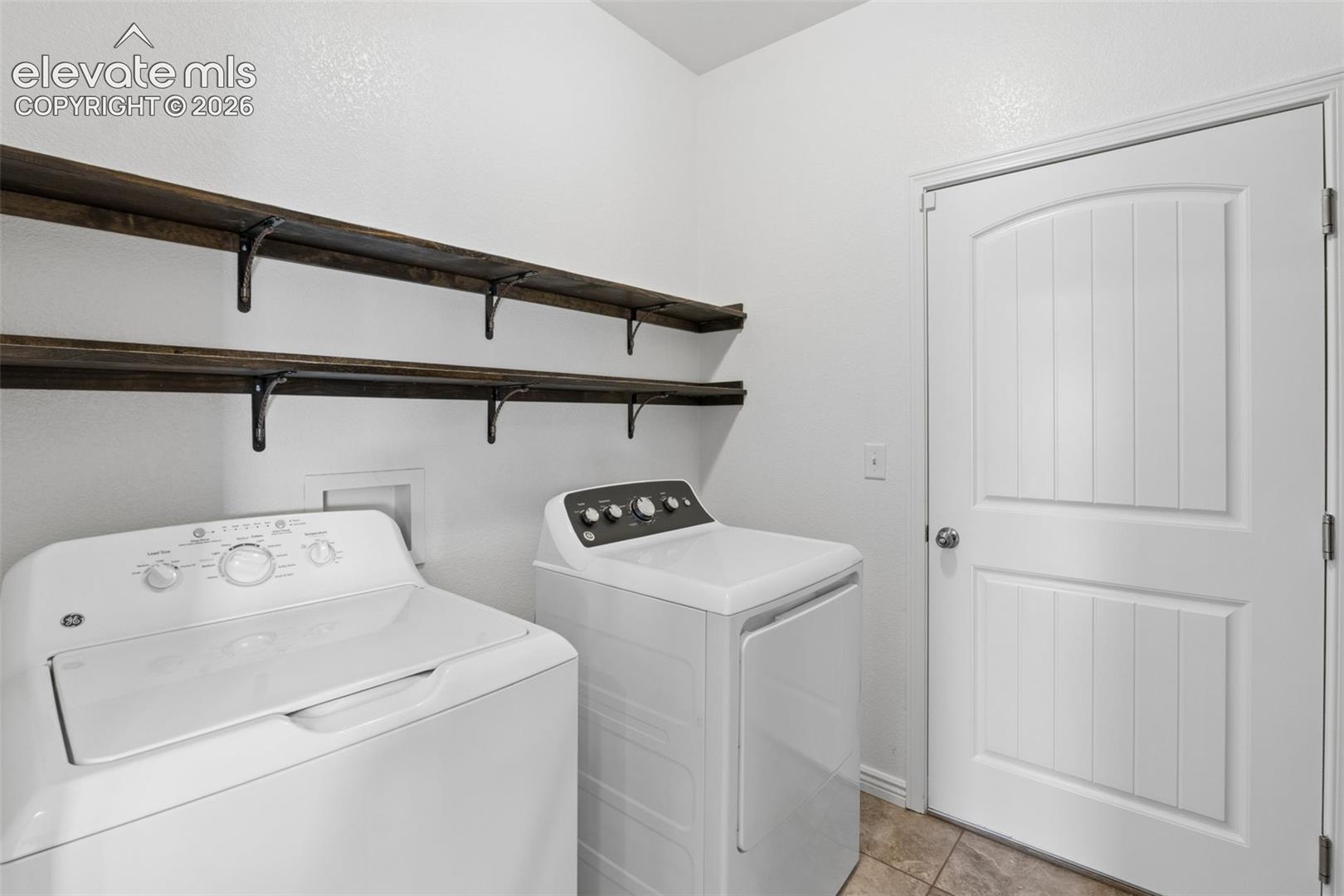 Dedicated laundry area featuring a white paneled door, two dark wood-finish floating shelves with metal brackets, and tile flooring. Leading to garage.