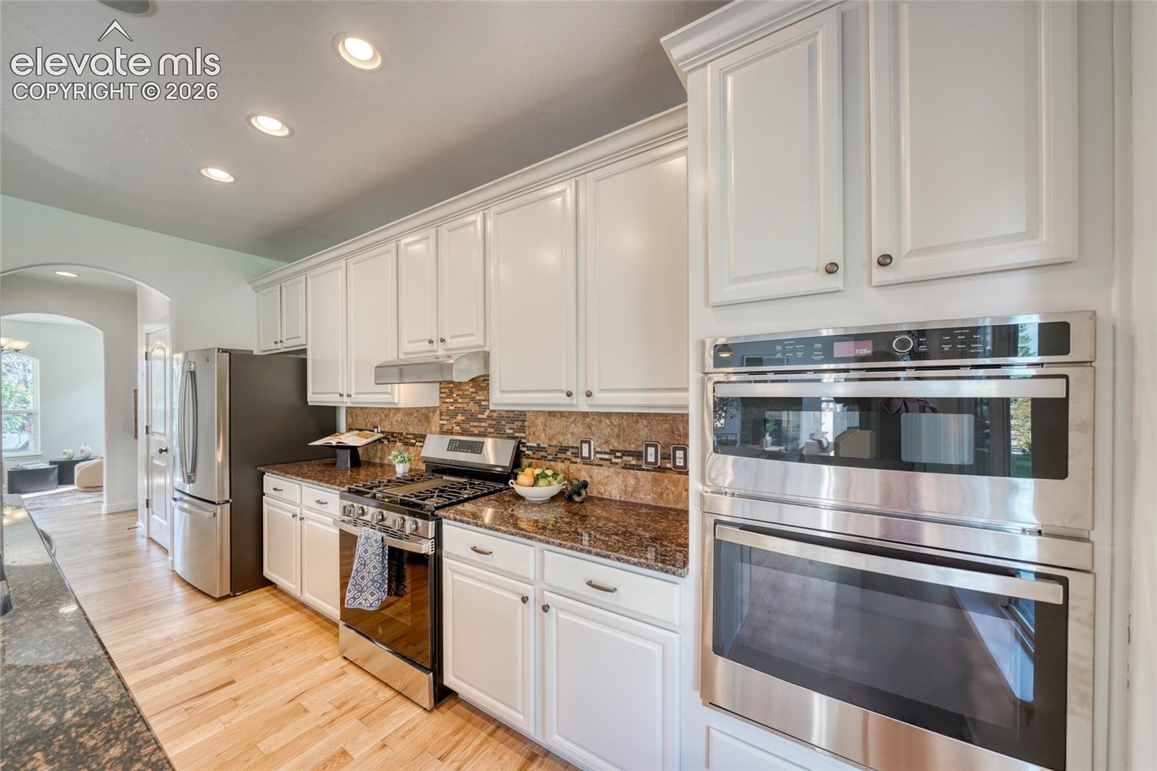 White cabinetry with crown molding, stainless steel appliances, a built-in oven and microwave, granite countertops, and wood-finish flooring