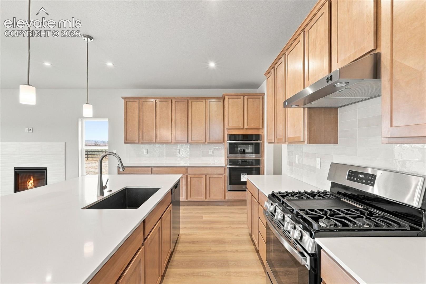 Kitchen featuring light wood-finish cabinetry, white countertops, stainless steel appliances including a double wall oven and gas range, subway tile backsplash, and two pendant lights