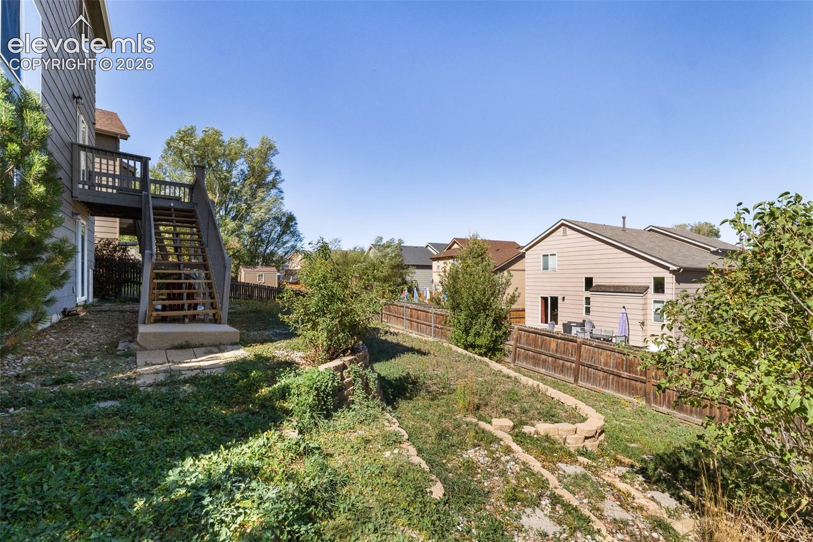 Rear exterior featuring a tiered stone retaining wall, wooden deck with staircase, and perimeter wood fencing