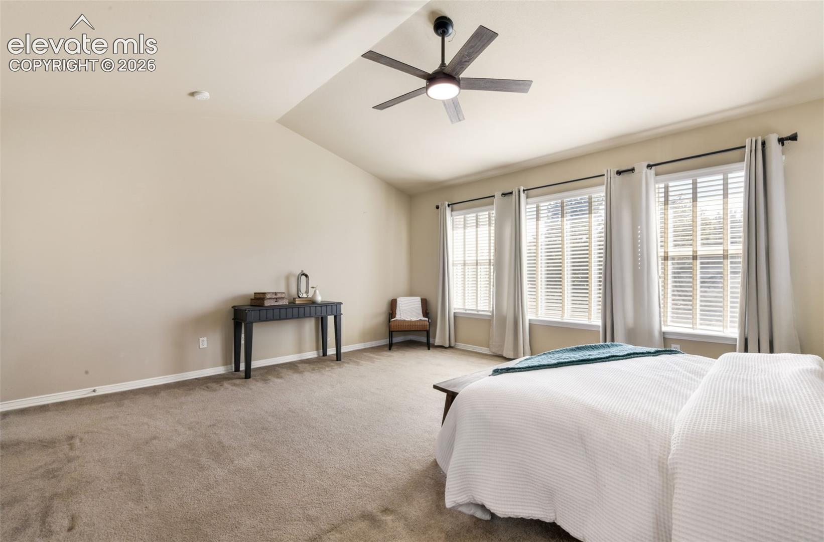 Vaulted ceiling with an attached ceiling fan, three large windows with blinds, and neutral-toned carpeting