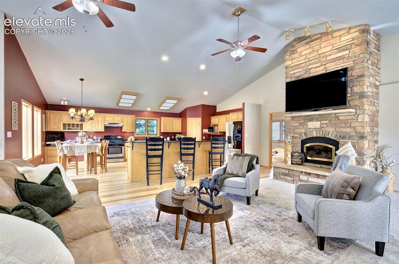 Vaulted ceilings featuring recessed lighting, dual ceiling fans, complemented by a floor-to-ceiling stone fireplace with a raised hearth, ash flooring, and cabinetry with an integrated breakfast bar
