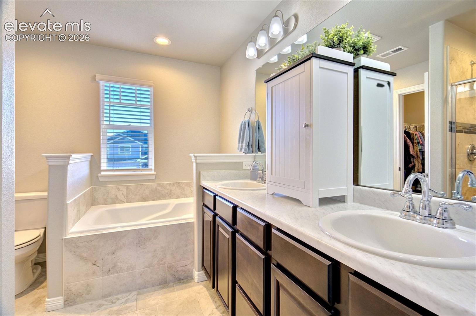 Dual vanity with dark cabinetry, stone-finish countertops, undermount sinks, tile-surround soaking tub, and wall-to-wall mirror