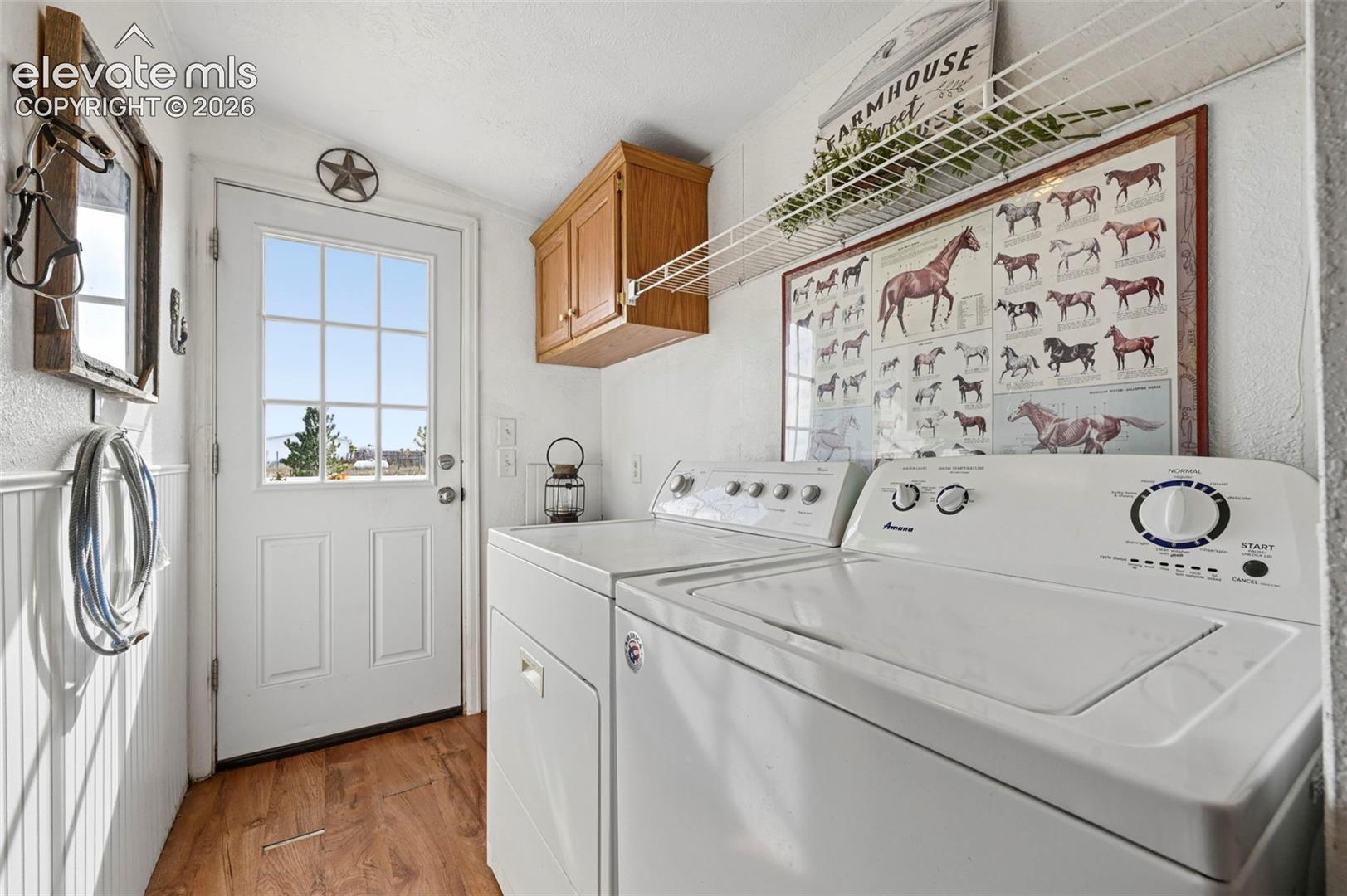 Laundry area featuring wood-finish flooring, white beadboard wainscoting, upper wood cabinetry, and an exterior door with a multi-pane glass insert