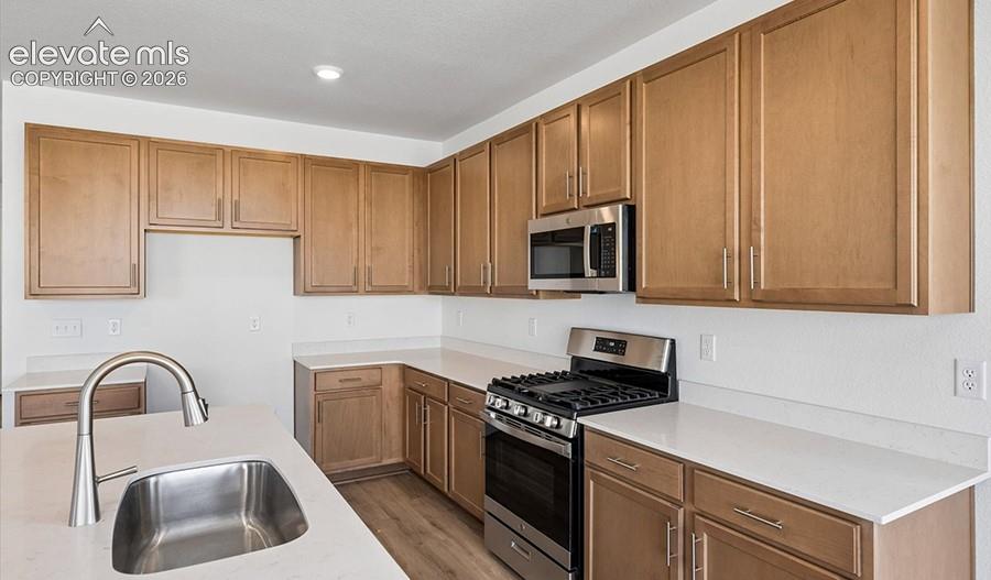 Kitchen featuring wood cabinetry, light-toned stone-finish countertops, stainless steel appliances, and a central island with an undermount sink and high-arc faucet