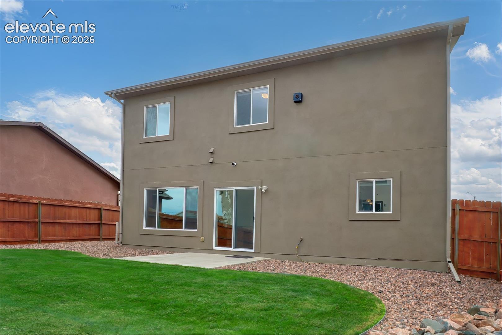 Two-story stucco exterior featuring a concrete patio, sliding glass door, fenced perimeter, and maintained lawn