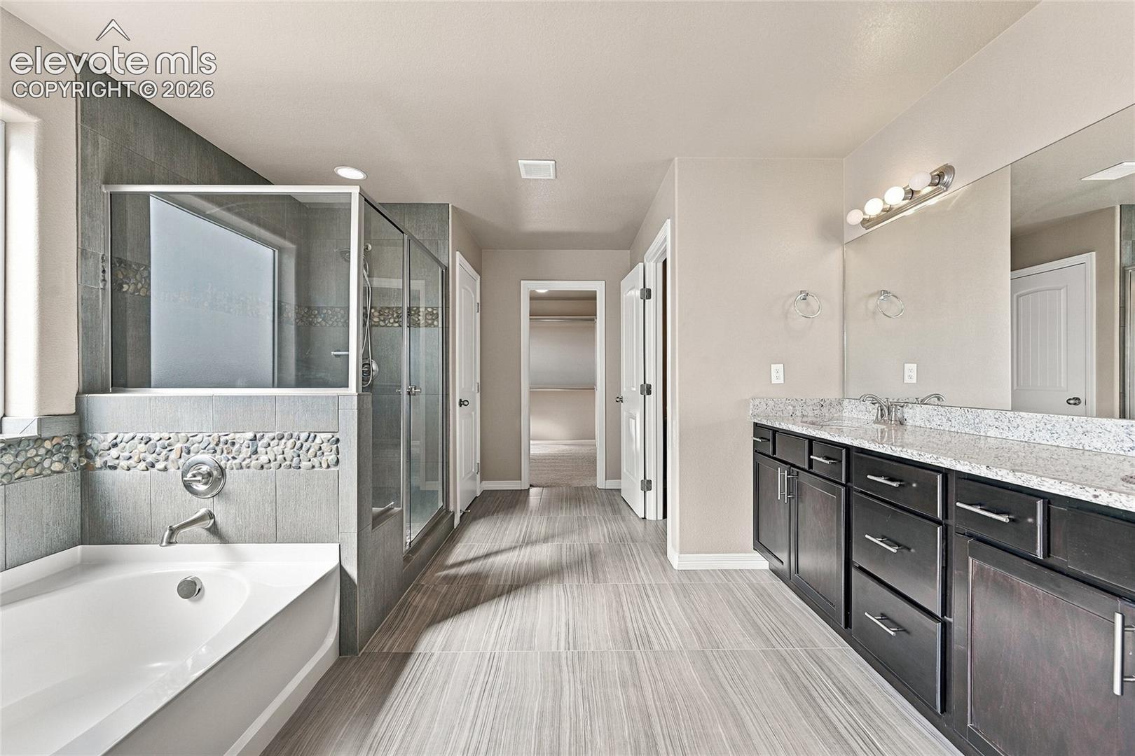 Bathroom featuring a deep-soaking tub, glass-enclosed shower with pebble tile accents, dual-sink vanity with stone-composite countertop, and wood-finish tile flooring