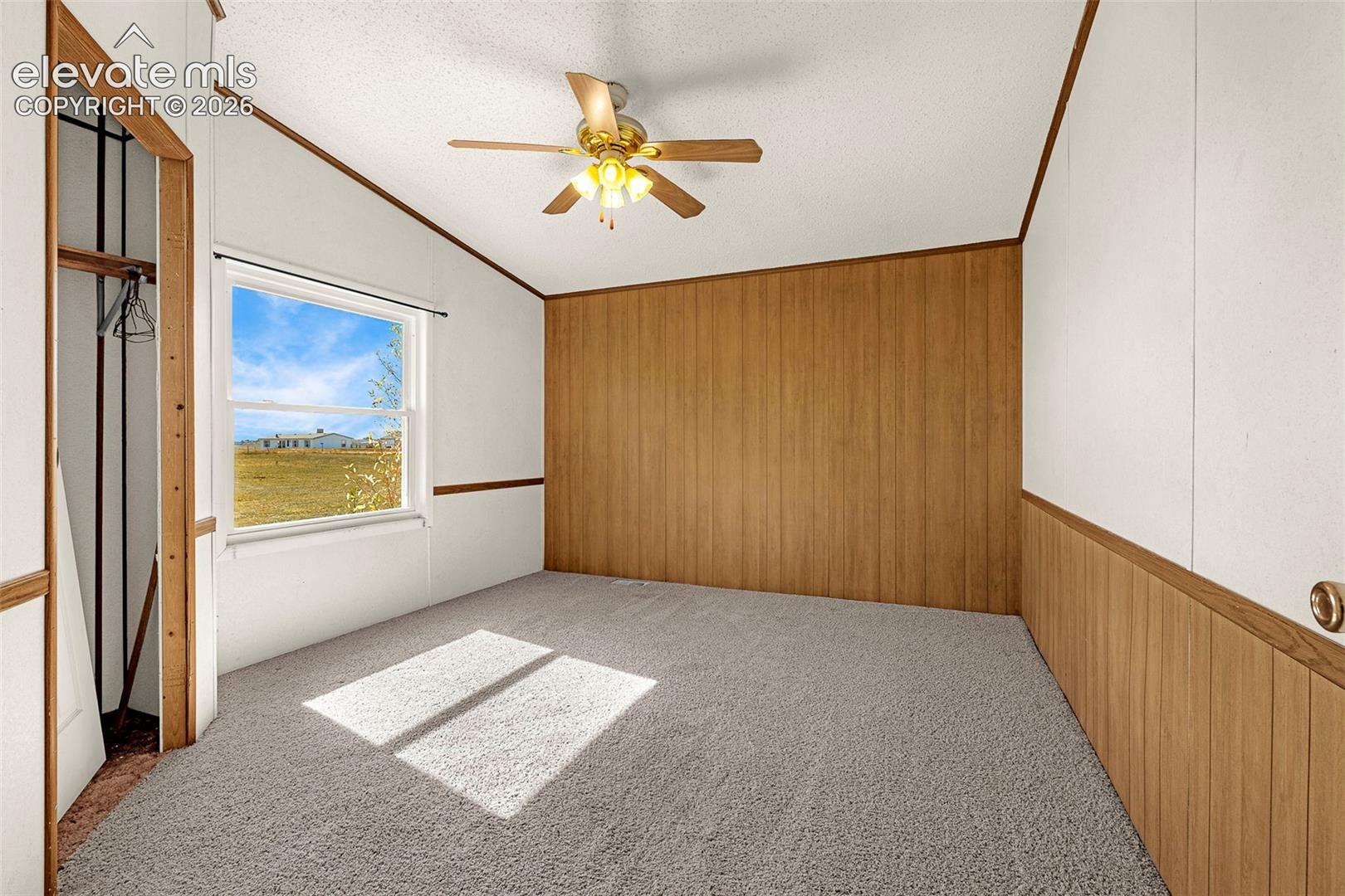 Vaulted ceiling featuring a central multi-blade ceiling fan, paneled wood accent walls, carpeted flooring, and a window with exterior landscape exposure