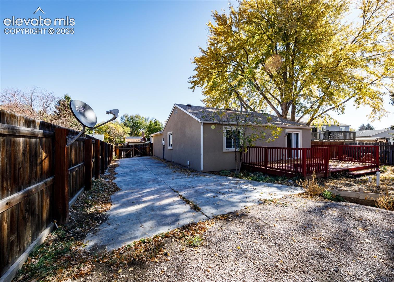 Concrete driveway, wood-plank perimeter fencing, raised wood deck, and mature deciduous tree