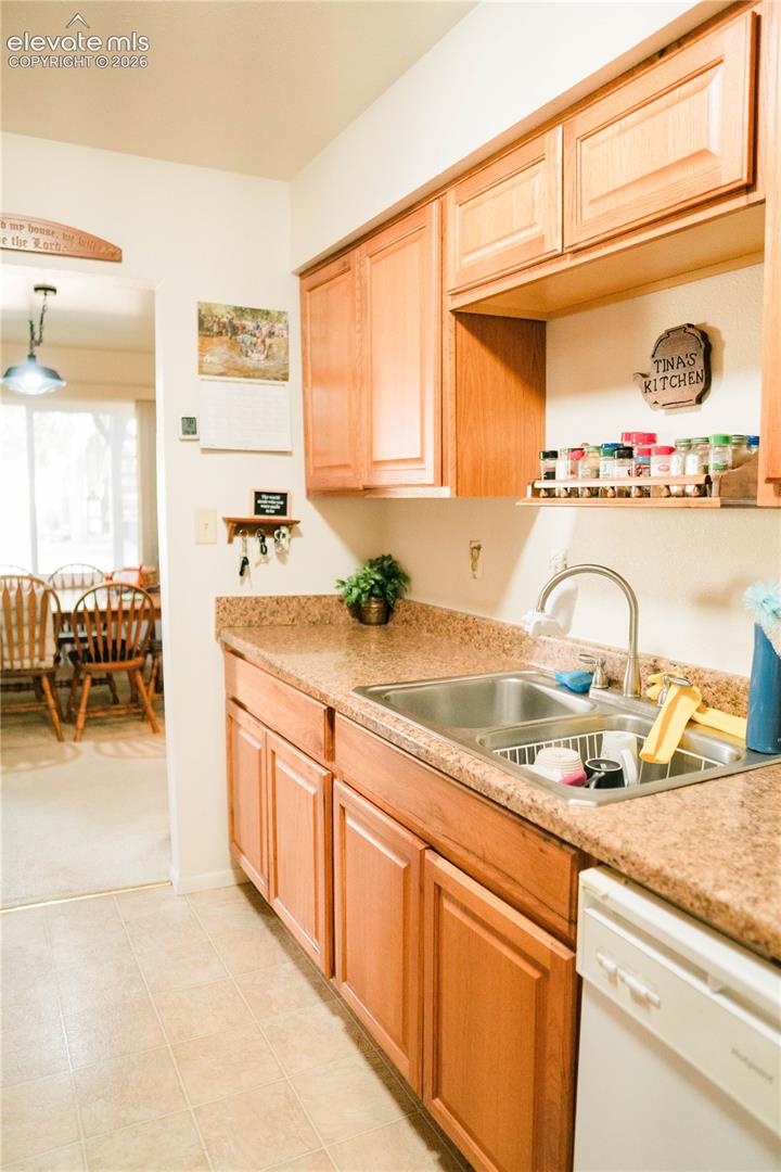Kitchen featuring wood-finish cabinetry, a double basin stainless steel sink with a gooseneck faucet, light-toned patterned countertops, and a built-in dishwasher