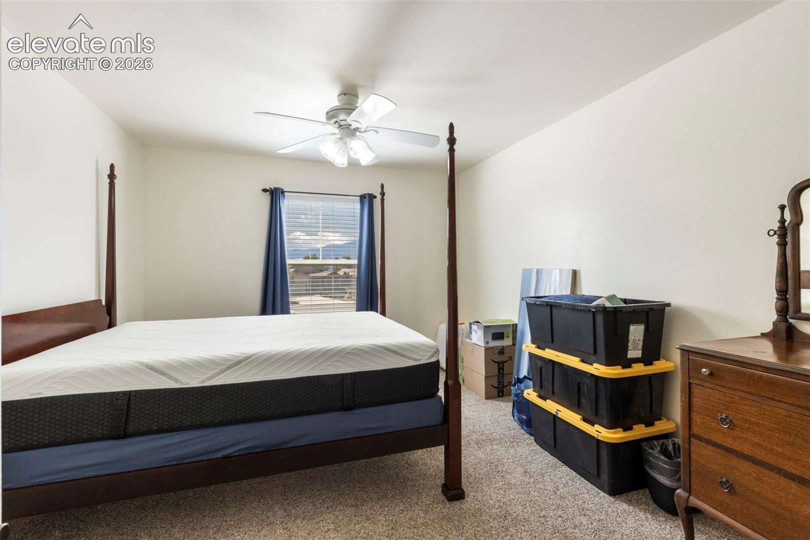 Spacious bedroom featuring a four-poster bed frame, ceiling fan with light fixture, carpeted flooring, and large window with blue drapery