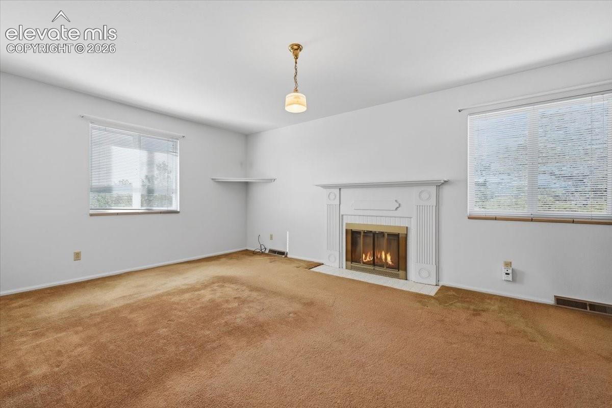 fireplace with a white millwork mantel, neutral wall-to-wall carpeting, two windows with integrated blinds, a built-in corner shelf, and a central pendant light fixture