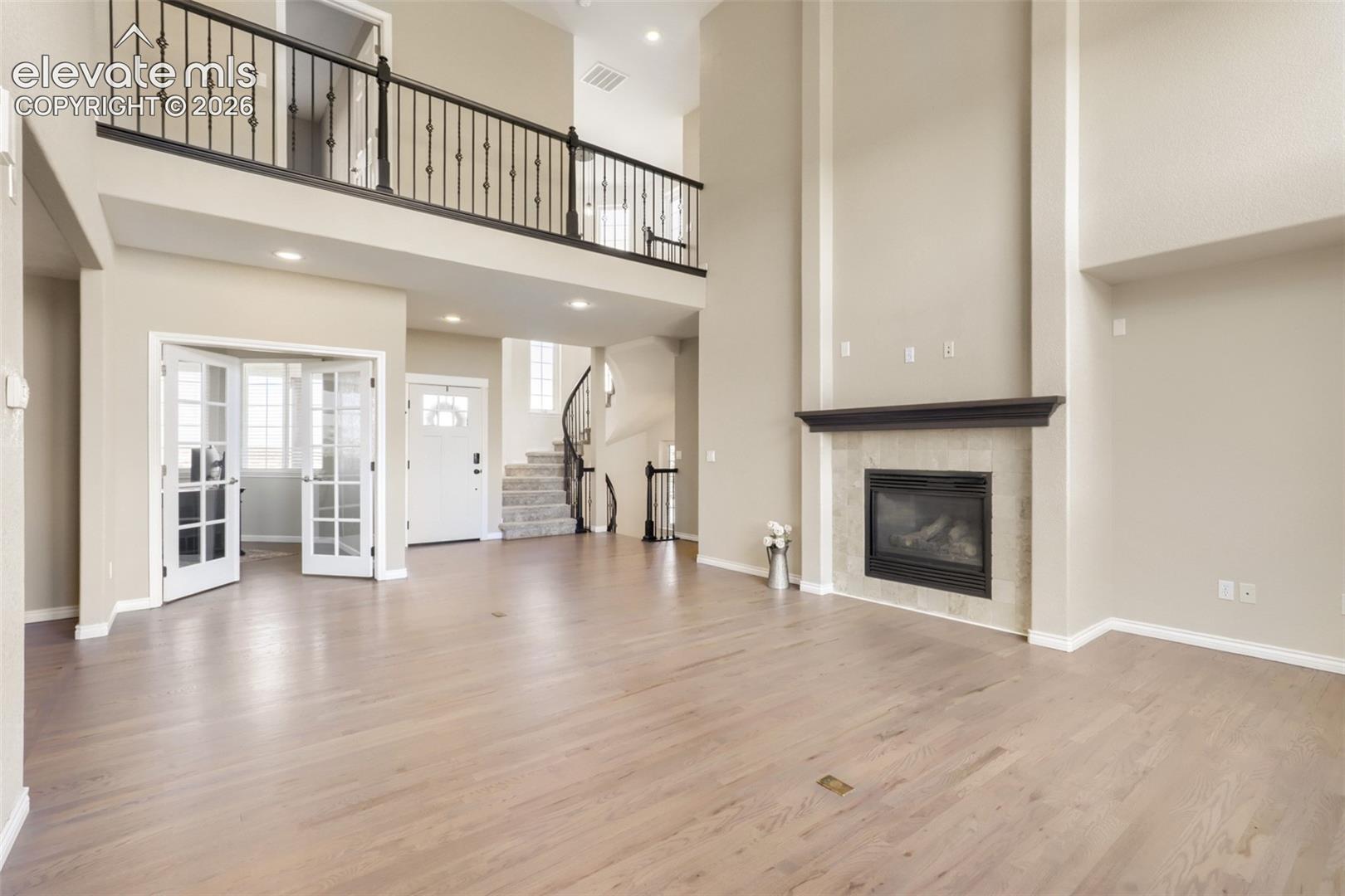 impressive two-story family room featuring a grand gas fireplace with tile surround, dark mantel, and floor-to-ceiling architectural framing. Overlooked by a second-story catwalk with wrought-iron balusters, this open layout seamlessly connects to the foyer, main staircase, and home office via glass French doors.