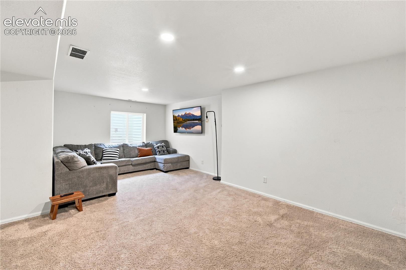 Recessed lighting and a single window with white horizontal blinds illuminate this carpeted room