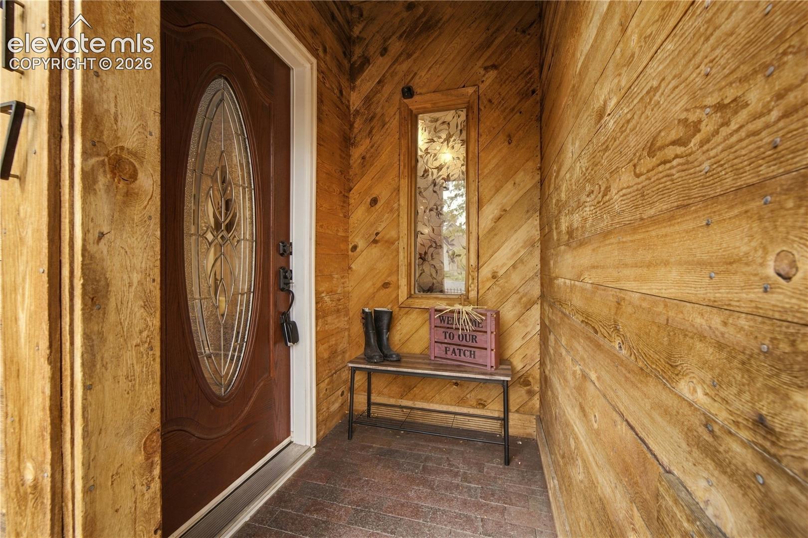 Entryway featuring wood-plank walls with diagonal and horizontal orientations, a dark wood-finish front door with an oval decorative glass insert, and brick-style flooring