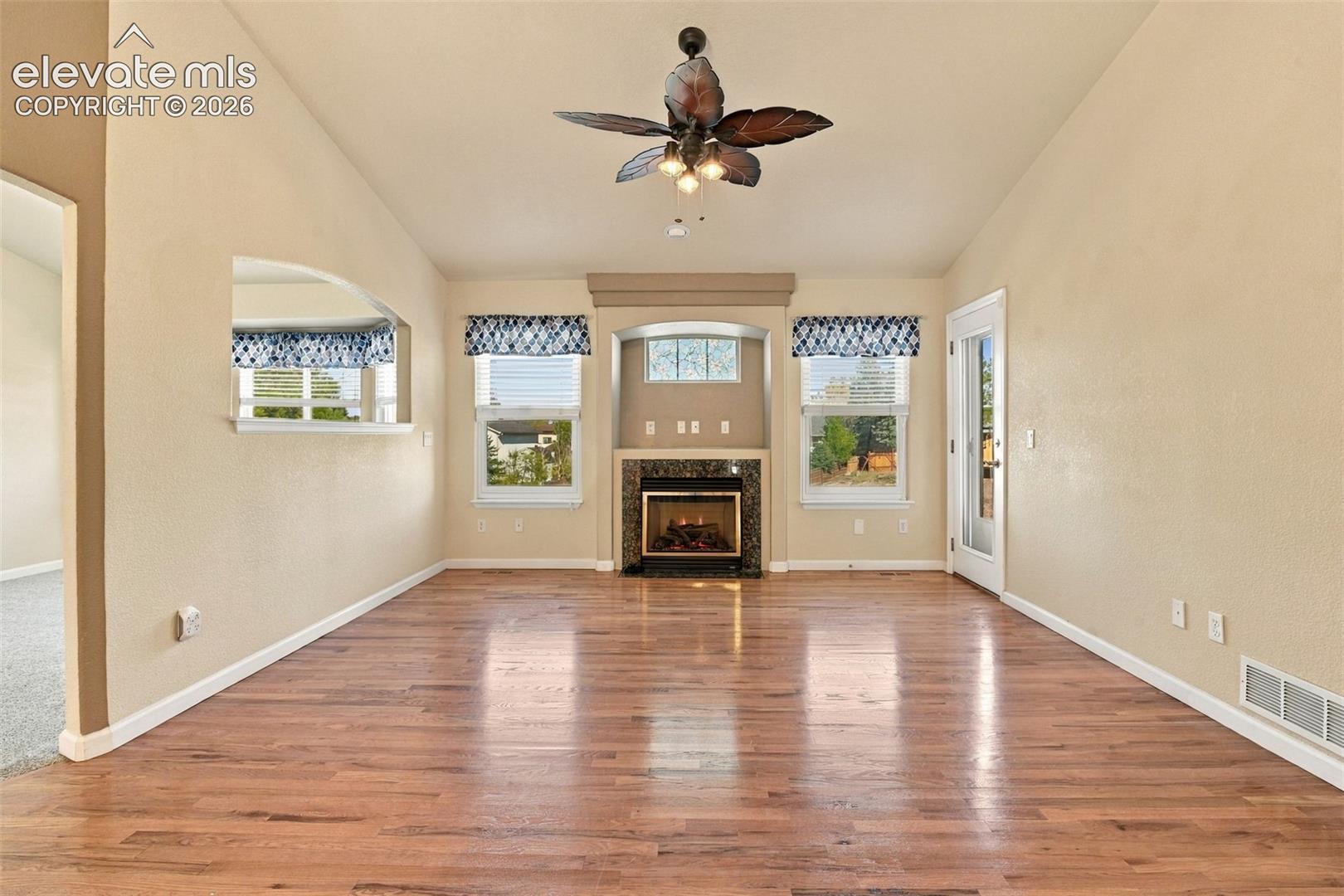 Living area featuring wood-finish flooring, a fireplace with a stone-surround mantel, vaulted ceilings, multi-pane windows, and a ceiling fan
