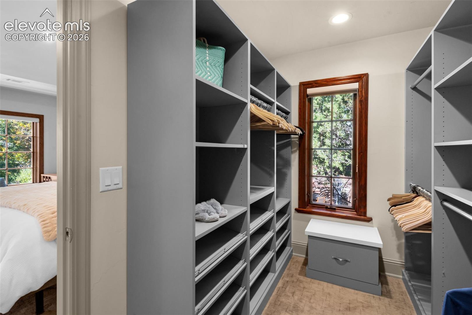 Custom built-in storage system featuring floor-to-ceiling shelving, integrated hanging rods, and a lower drawer unit with a solid surface top, accented by a window with stained wood trim and recessed lighting