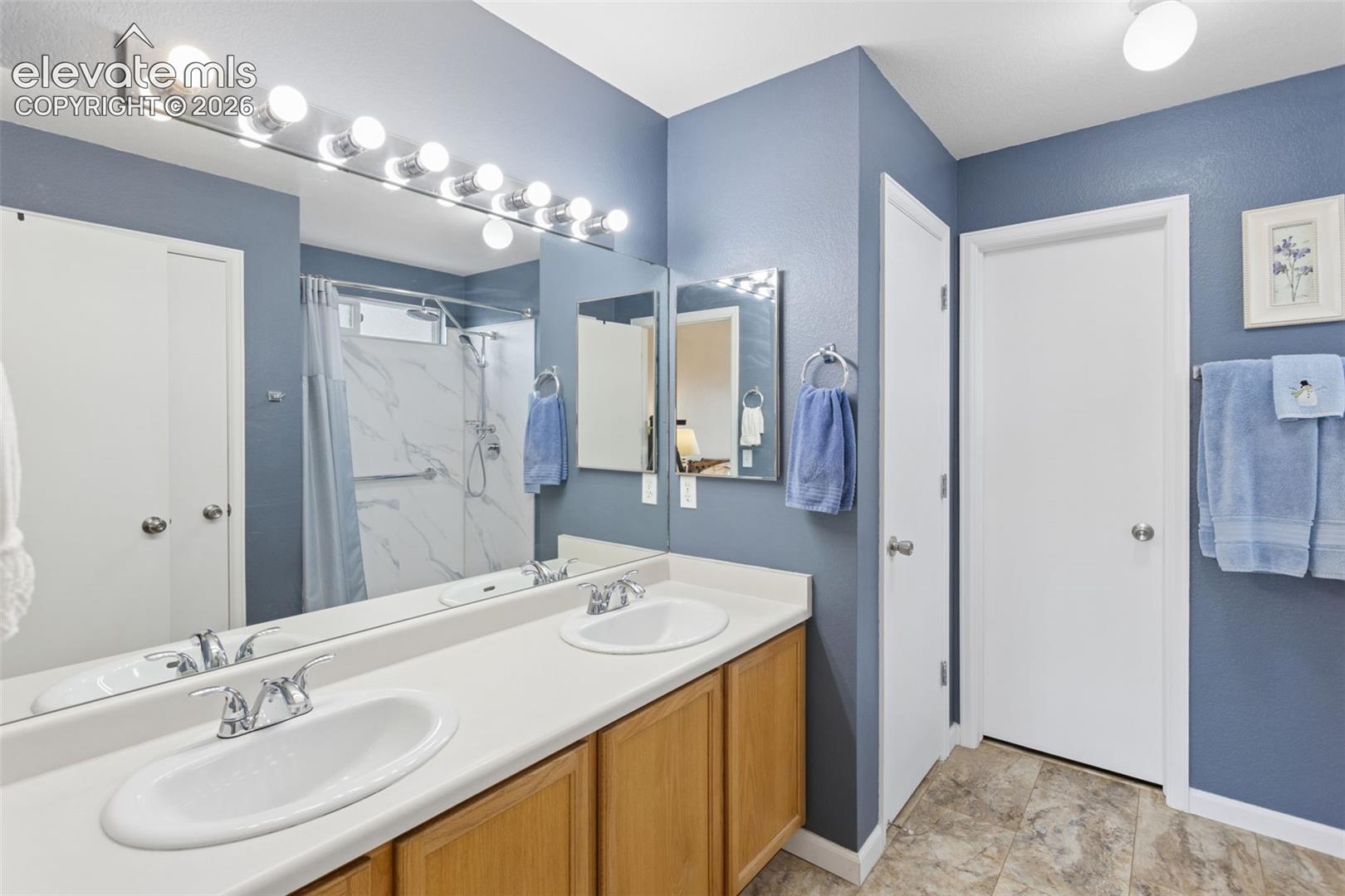 Dual sinks set in a white countertop, wood-finish cabinetry, a large vanity mirror with strip lighting, a shower with marble-look wall surround, and tile flooring
