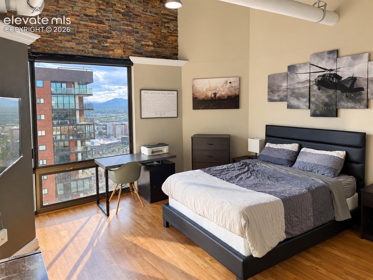 Expansive window wall, wood-finish flooring, exposed brick accent wall, high ceilings, and visible ductwork