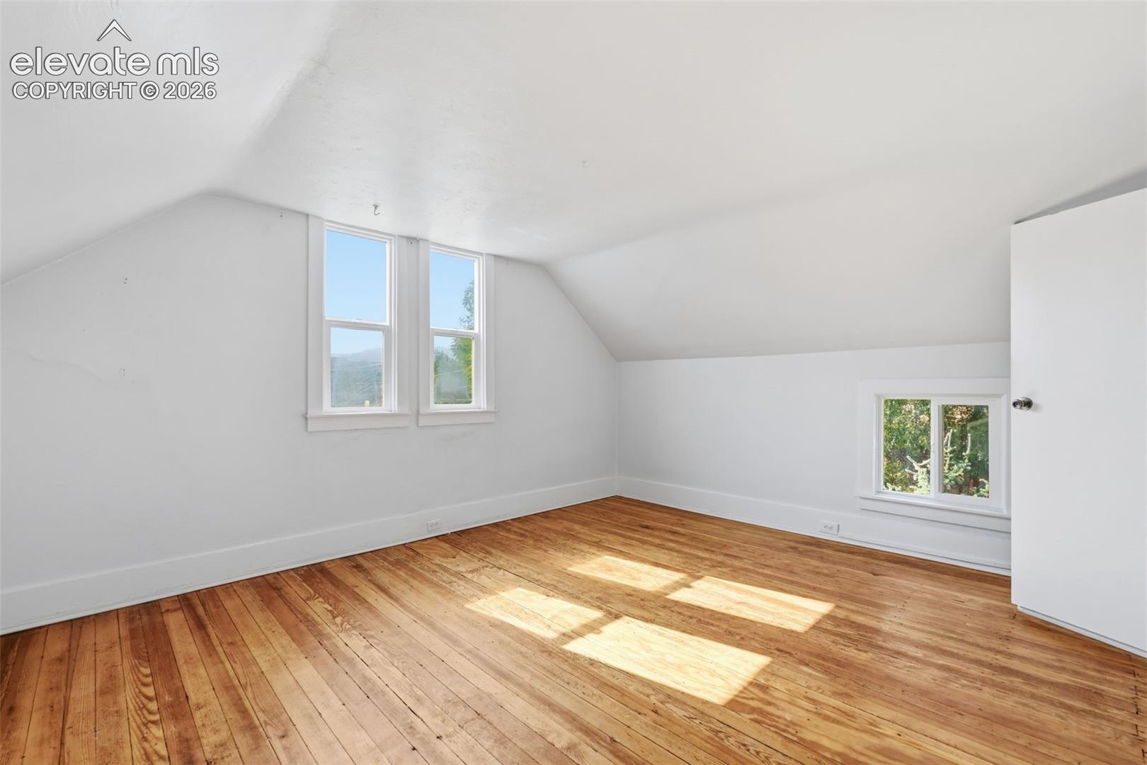 Primary bedroom with sloped ceilings, pine hardwood flooring, two large windows, and a smaller window.