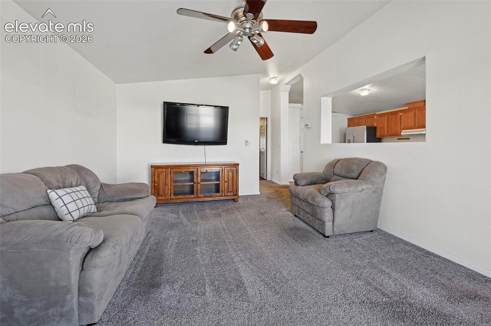 Living area featuring neutral carpeting, a ceiling fan with multi-light fixture, and an open wall cutout providing a sightline into the kitchen with wood cabinetry