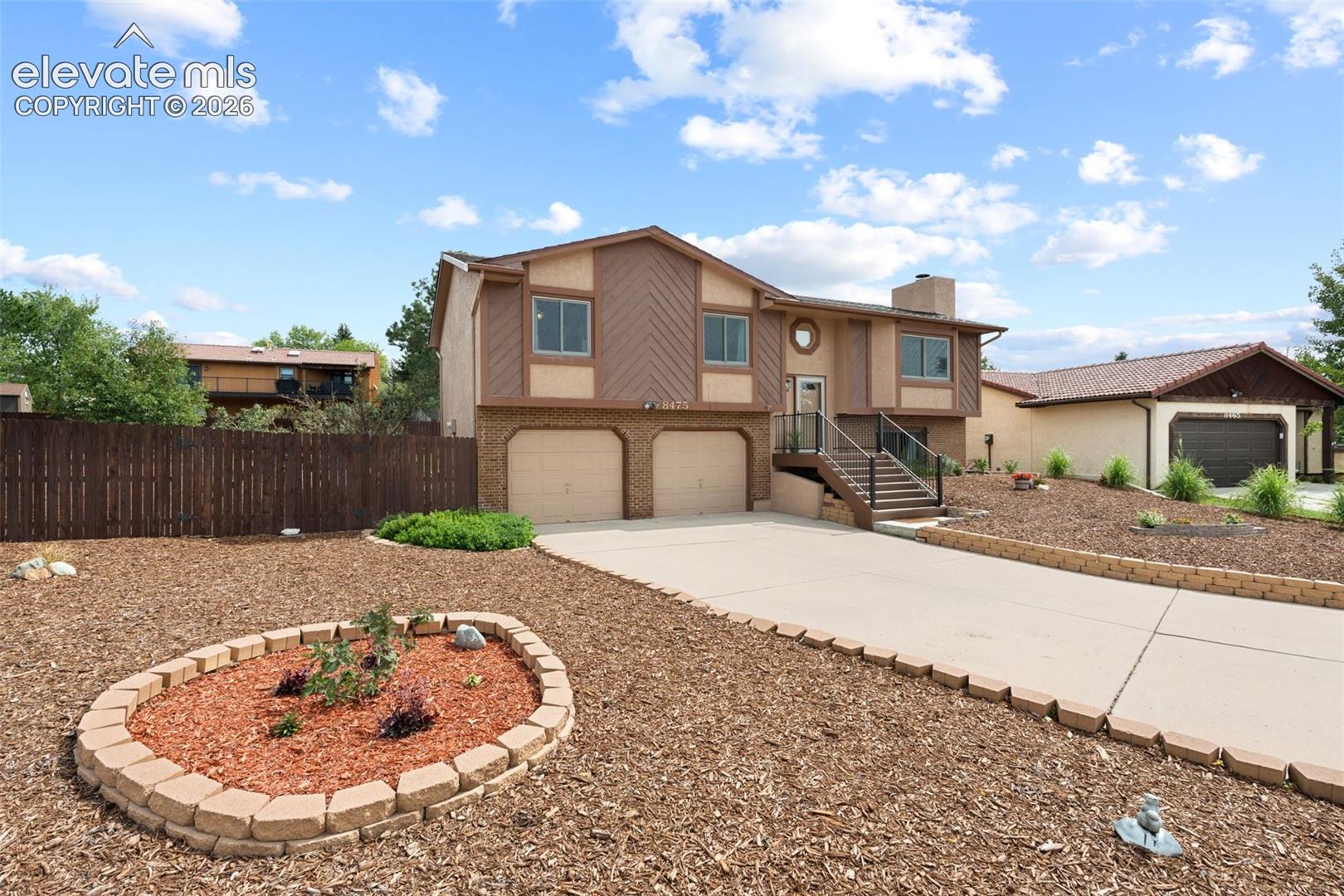 Multi-level exterior featuring a two-car garage, concrete driveway, elevated entryway with metal railing, and stone-block landscaping borders