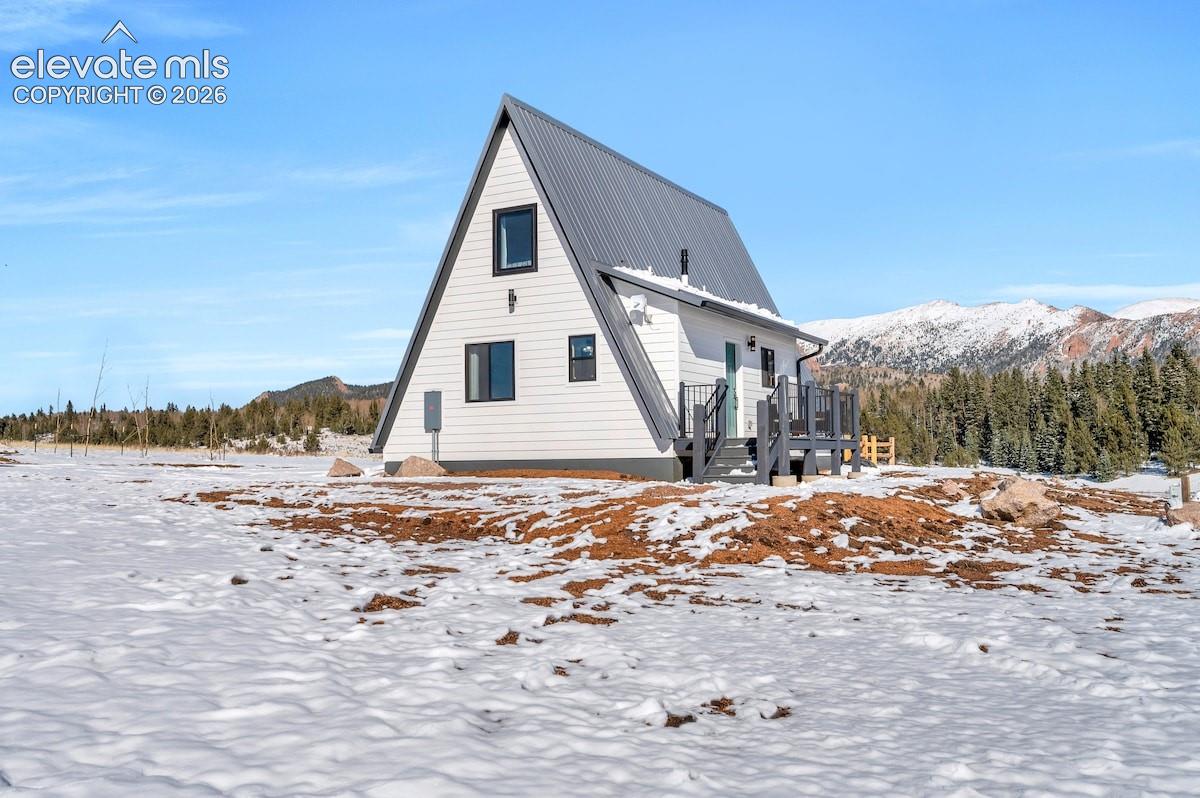 A-frame exterior featuring white horizontal siding, a charcoal metal roof, and an attached deck with metal railings, set against a backdrop of mountain peaks