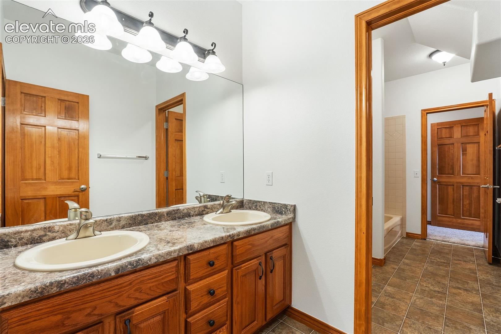 Double vanity featuring stone-finish countertops, dual integrated oval sinks, wooden cabinetry, and a full-width mirror with integrated vanity lighting