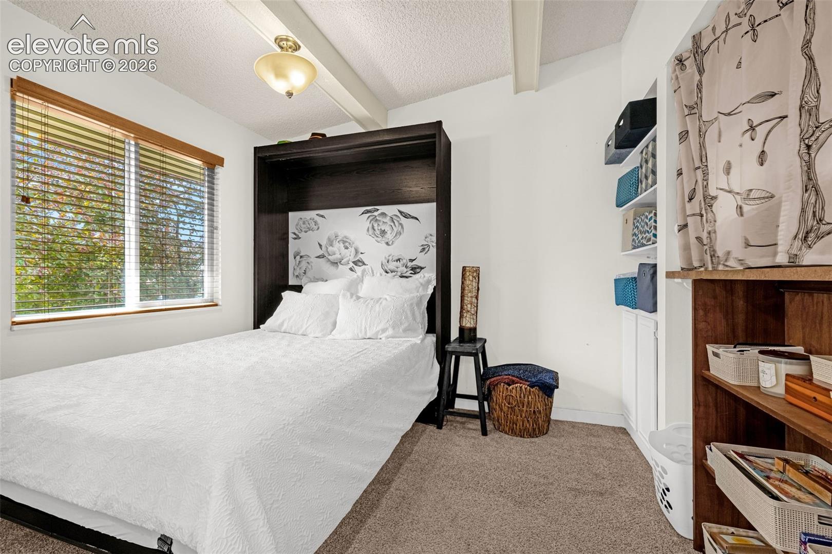 Angled vaulted ceiling with exposed beams, built-in murphy bed, built-in shelving, carpeted flooring, and a large window with blinds