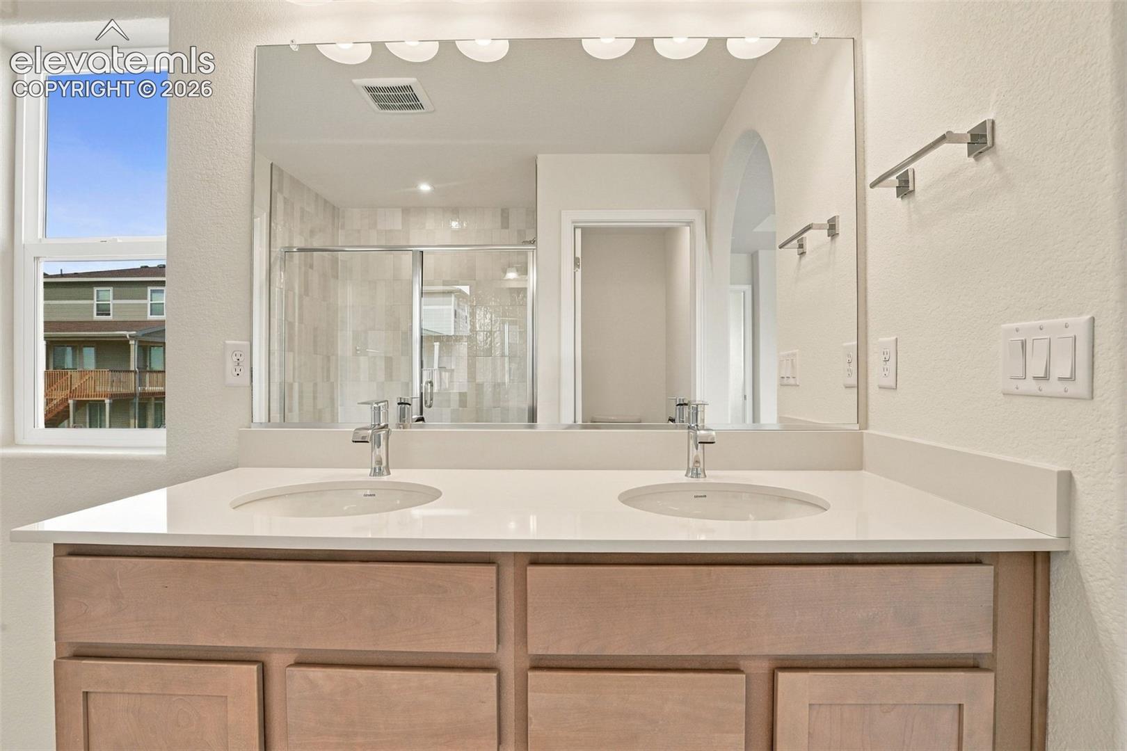 Double vanity featuring light wood-finish cabinetry, a solid surface countertop, integrated dual basins, polished chrome fixtures, and an expansive wall-mounted mirror with integrated lighting