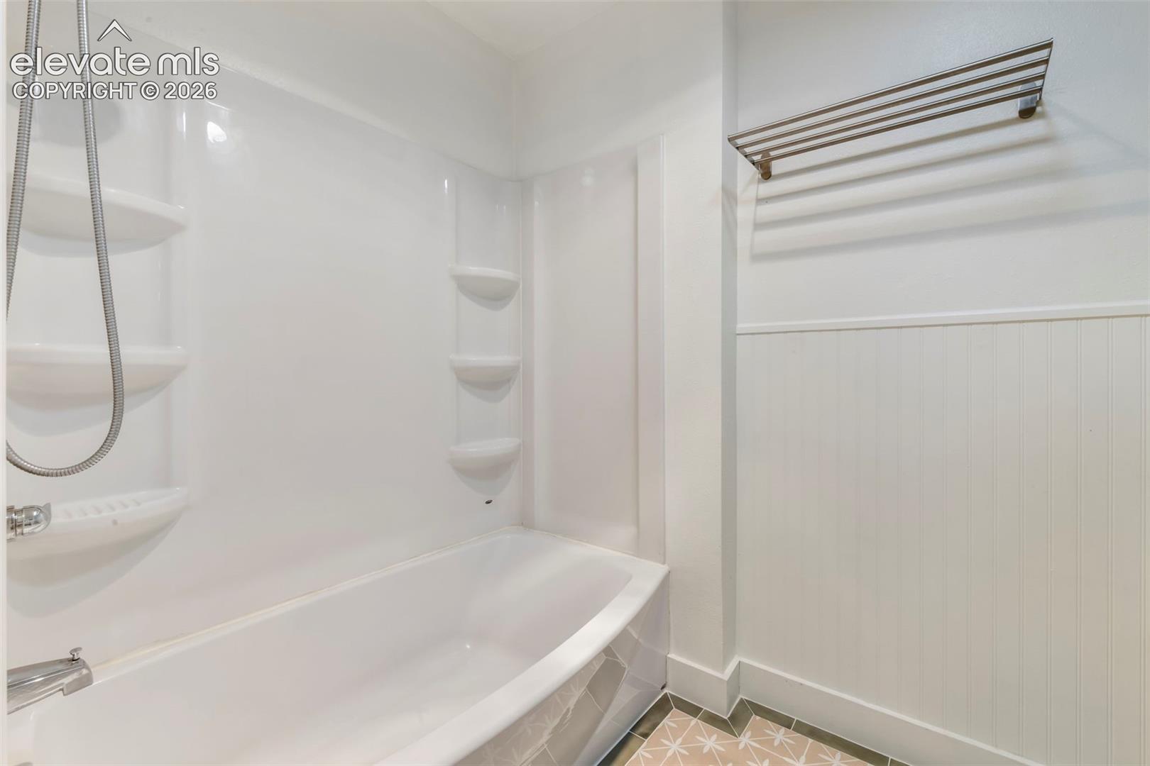 Bathtub featuring a molded surround with integrated shelving, a handheld shower fixture, beadboard wainscoting, and a wall-mounted towel rack