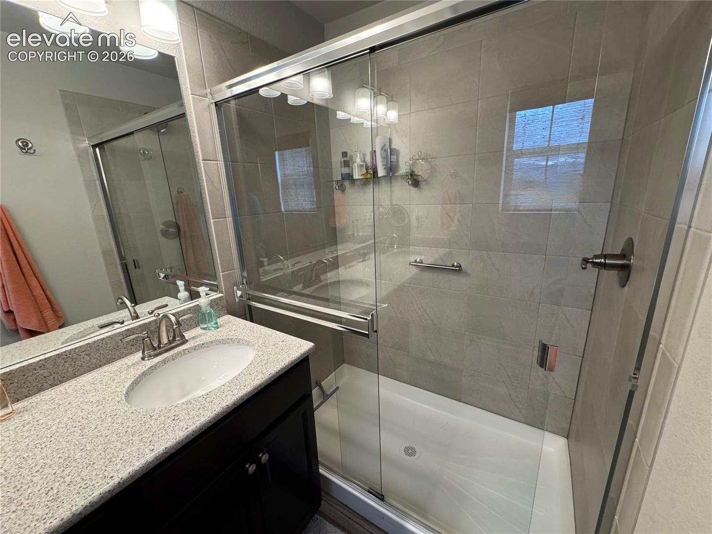Dark cabinetry with a light-toned countertop, oval undermount sink, large wall-mounted mirror, and a walk-in shower enclosure featuring floor-to-ceiling tile surround and sliding glass doors