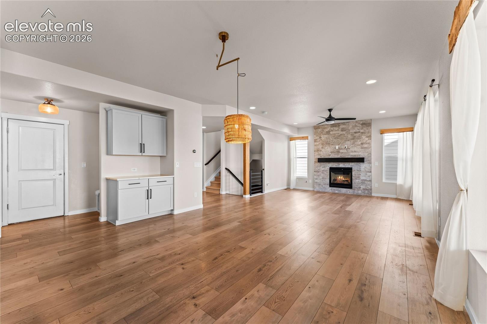 Open-concept living space featuring wood-finish flooring, a full-height stone fireplace surround with a linear mantel, built-in cabinetry with light-toned counters, and a hanging woven pendant fixture