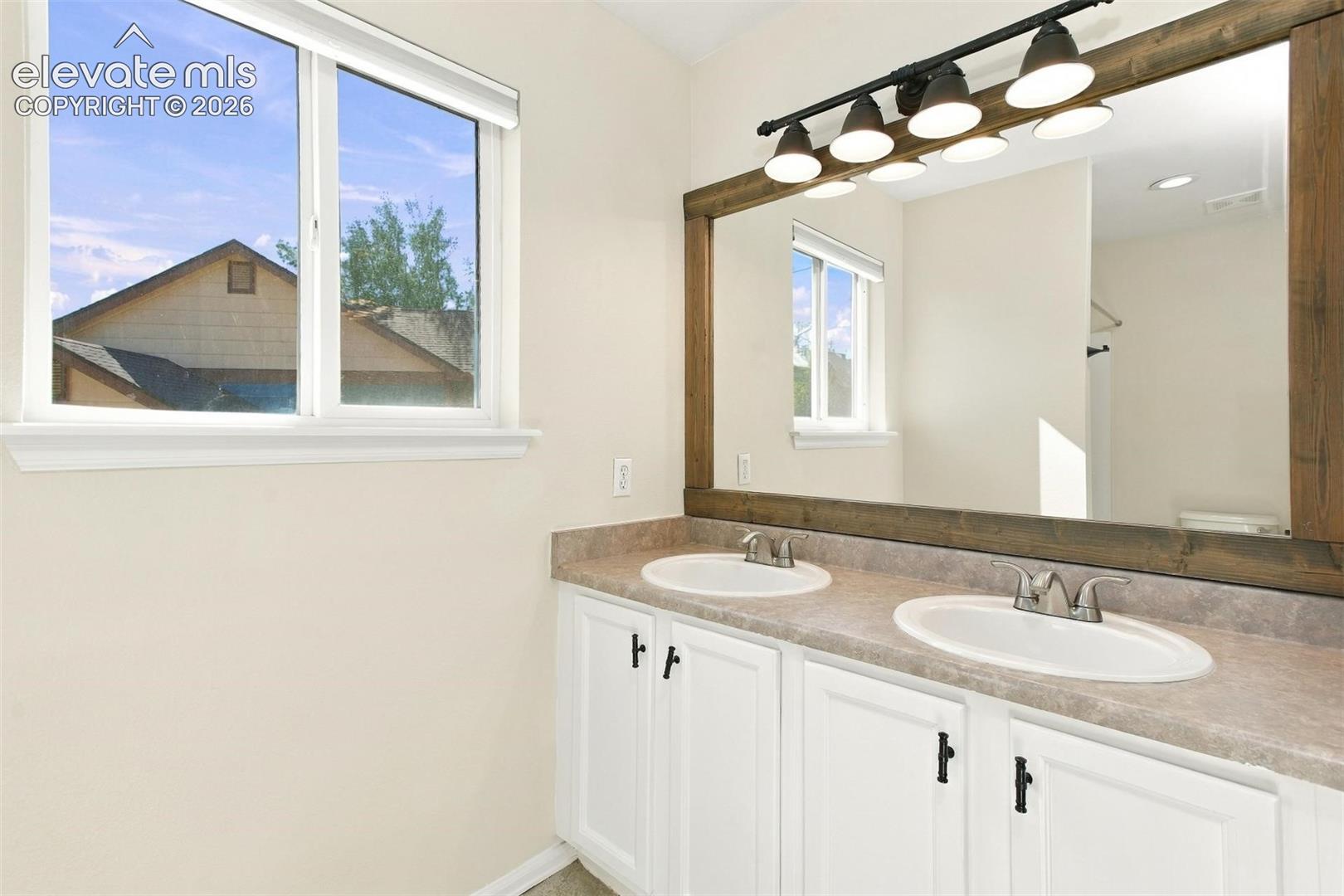 Dual-sink vanity with a stone-finish countertop, white cabinetry with black hardware, large mirror with a wooden frame, and multi-bulb integrated lighting fixture