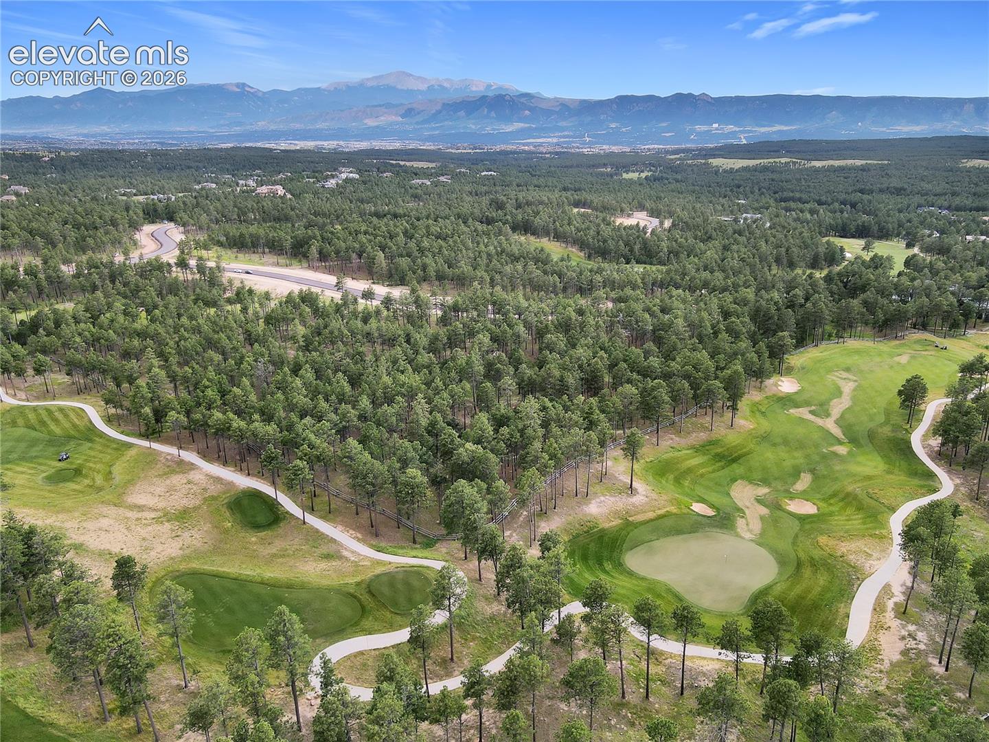 Aerial landscape featuring a manicured golf course, dense evergreen forest, winding paved paths, and prominent mountain backdrop