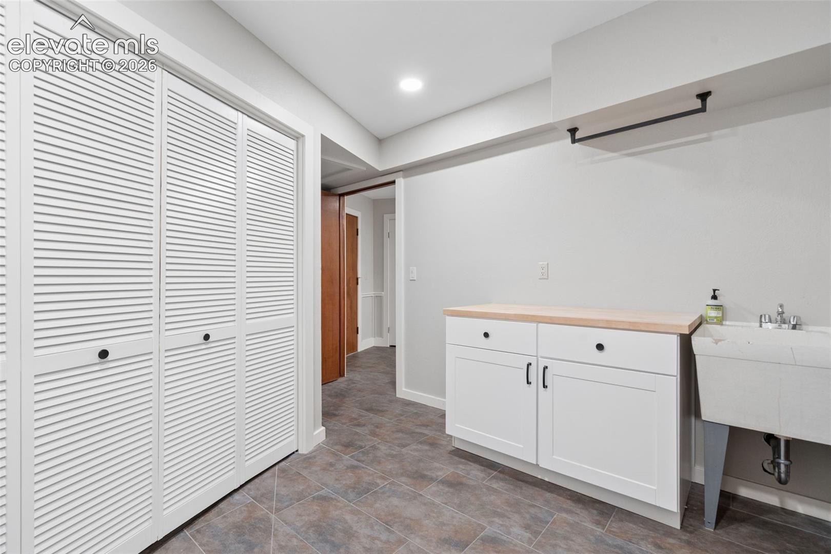 White louvered bi-fold doors, recessed lighting, tile flooring, white cabinetry with wood-finish countertop, and a utility sink