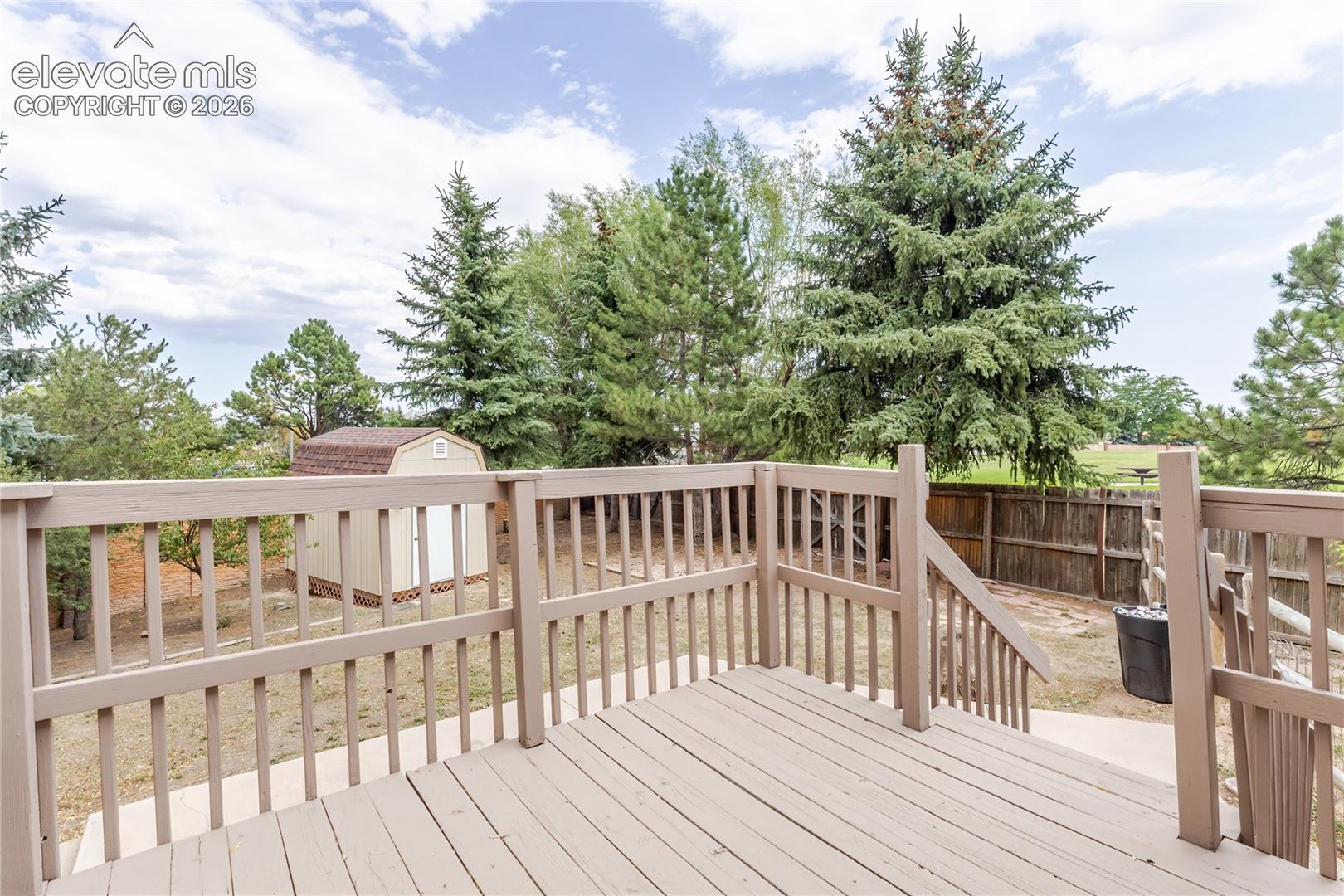 Wood-finish deck with railing and integrated steps, featuring an exterior shed and mature trees