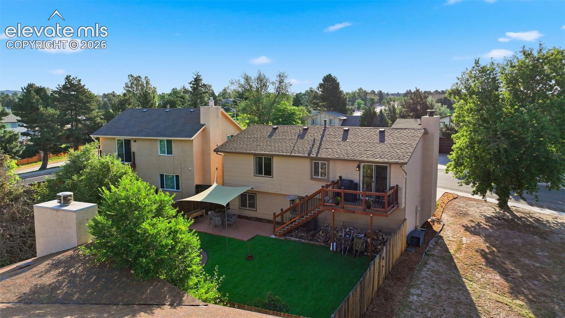 Rear exterior featuring a multi-level deck with wooden railing and stairs, a patio with sunshade, a fenced yard with artificial turf, and mature landscaping