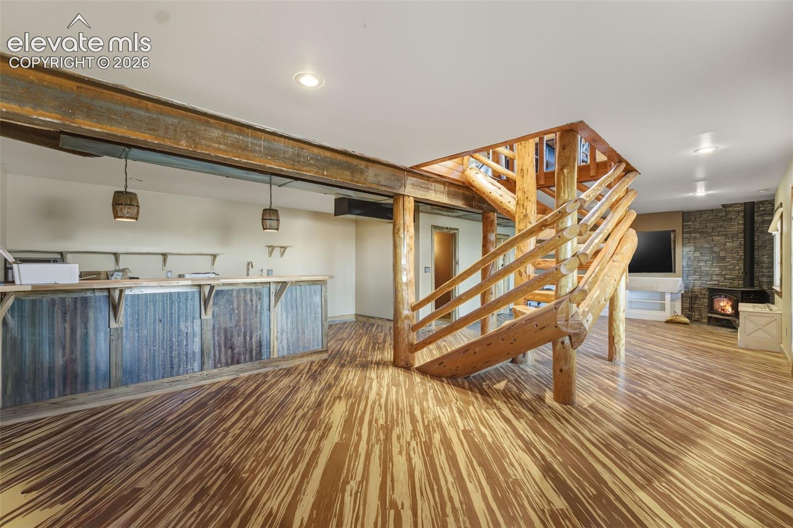Custom wood flooring, wood beam accents, built-in bar with corrugated metal and wood paneling, log staircase, and a stone-clad wall with a freestanding stove