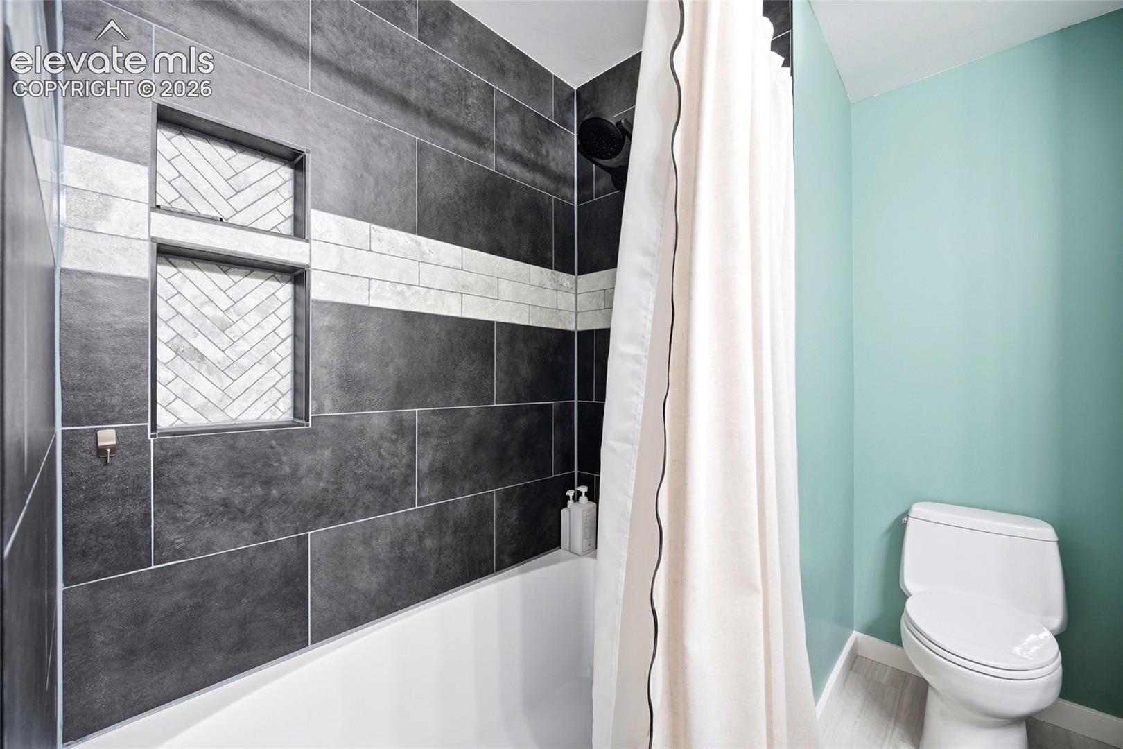 Tub and shower combination featuring dark large-format tile surround, integrated herringbone-patterned recessed shelving, horizontal light-toned accent tiling, and matte black shower fixtures