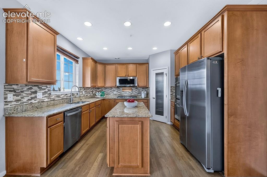 Wood-finish flooring, recessed lighting, wood cabinetry, stainless steel appliances, and a central island with a granite-style countertop
