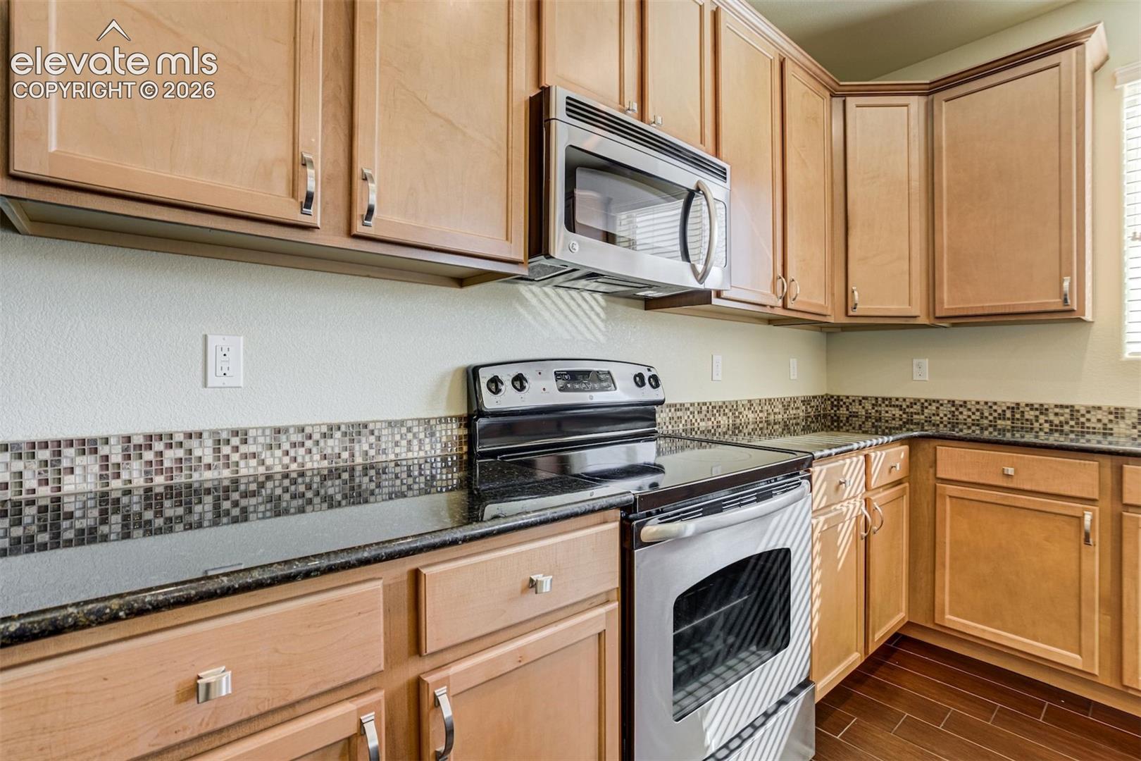 Wood-finish cabinetry with silver-tone hardware, stainless steel microwave, black electric range with stainless steel oven, dark countertop, and mosaic tile backsplash