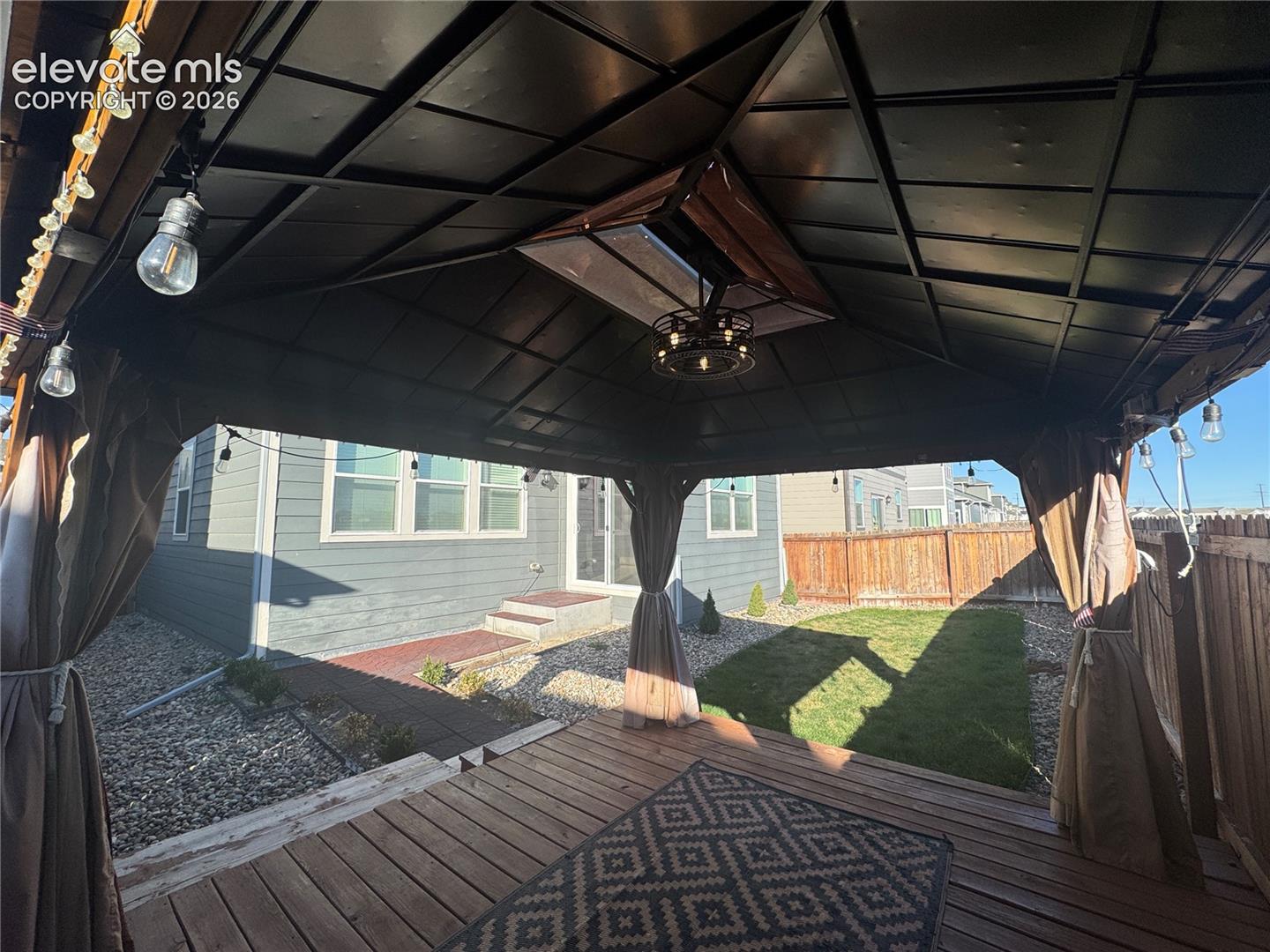 Covered wooden deck featuring a metal-frame gazebo, integrated ceiling lighting, and hardscaped brick pathing bordering a fenced lawn