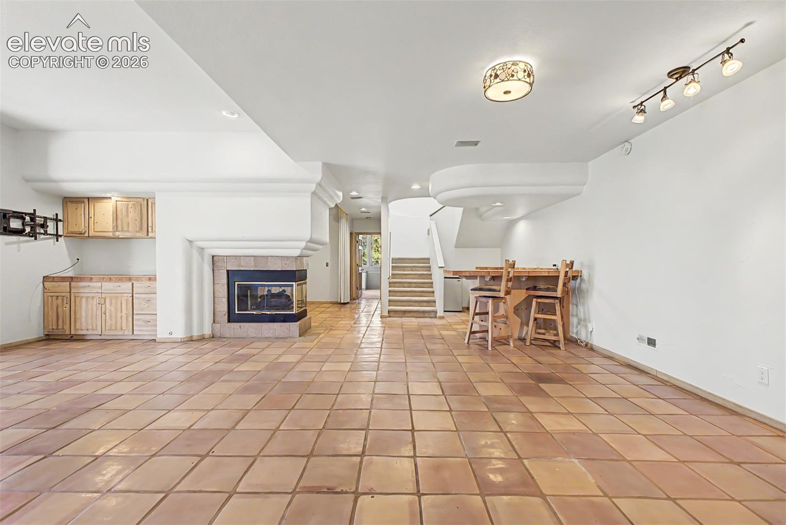 Terracotta tile flooring, built-in wet bar cabinetry, corner fireplace with hearth, and staircase